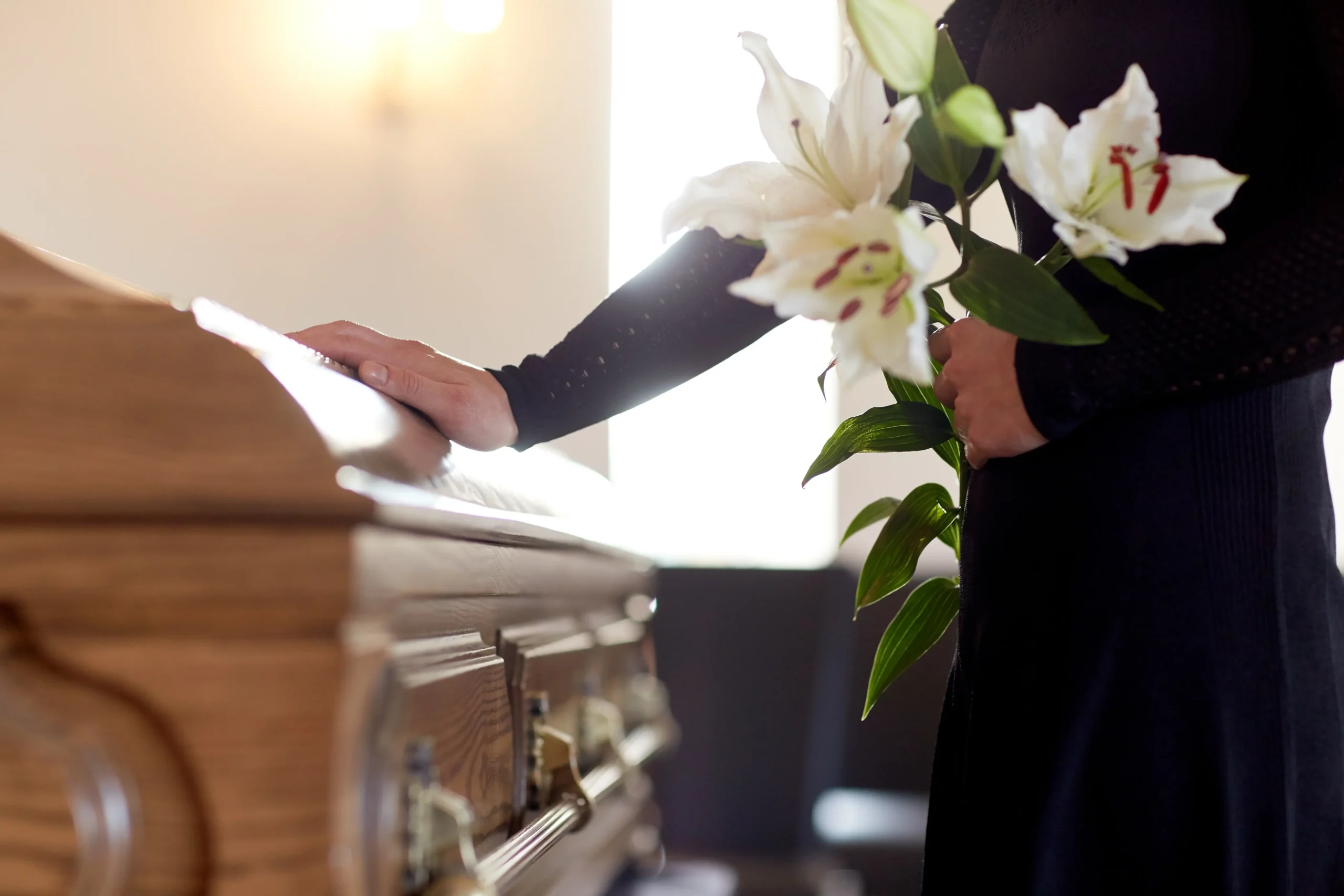 A person in dark clothing resting a hand on a wooden casket while holding white lilies