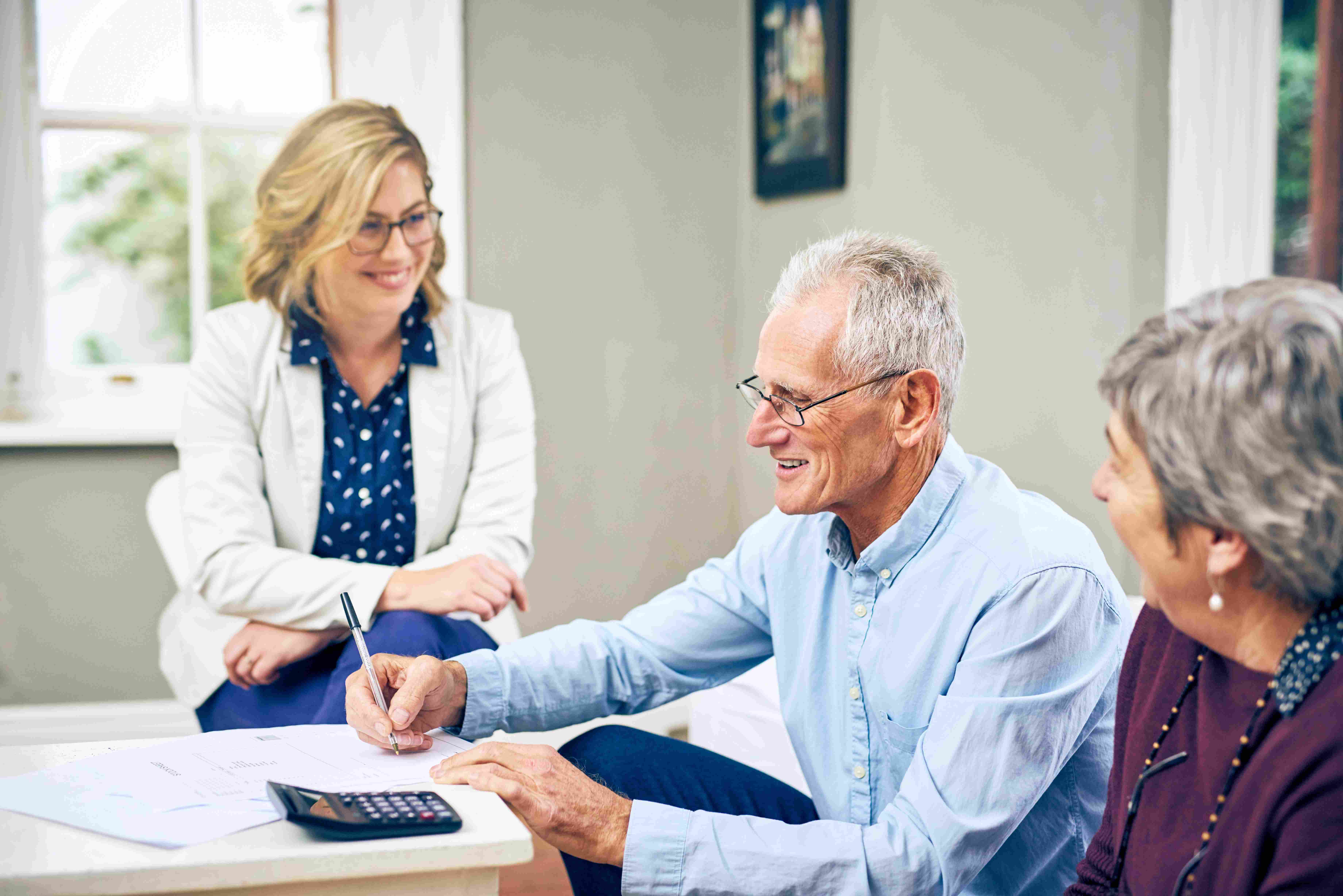 An older couple signing paperwork at a table with a consultant and a calculator nearby