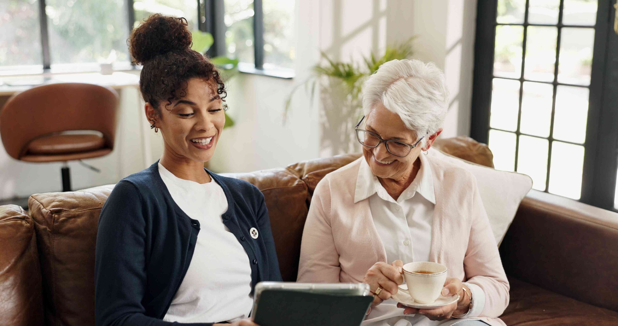 Two women sitting on a couch together, smiling while looking at a tablet, one holding a cup of tea